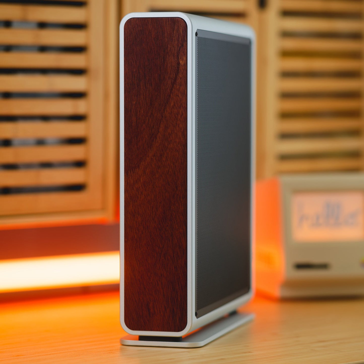 A modern Computer with a dark wood grain front and silver side trim, standing on a small base on a wooden table with warm ambient lighting and a blurred vintage-style clock in the background.